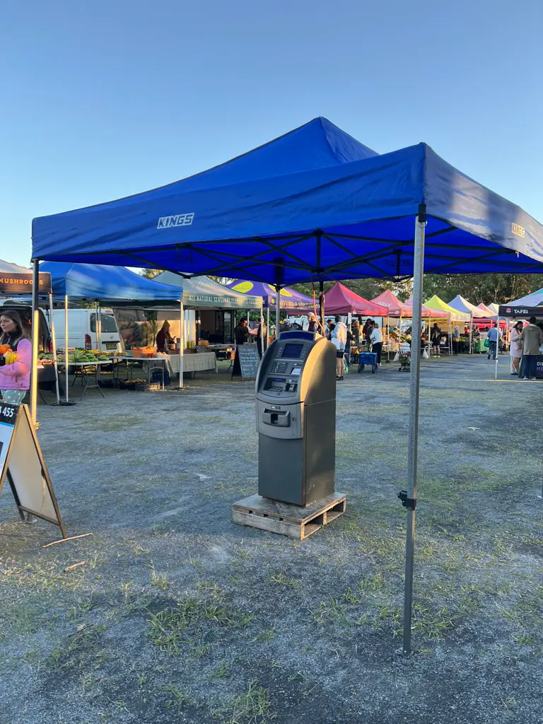 A market ATM under a blue gazebo at dawn with producer stalls and a frosty field behind at the Byron Bay Farmers Market