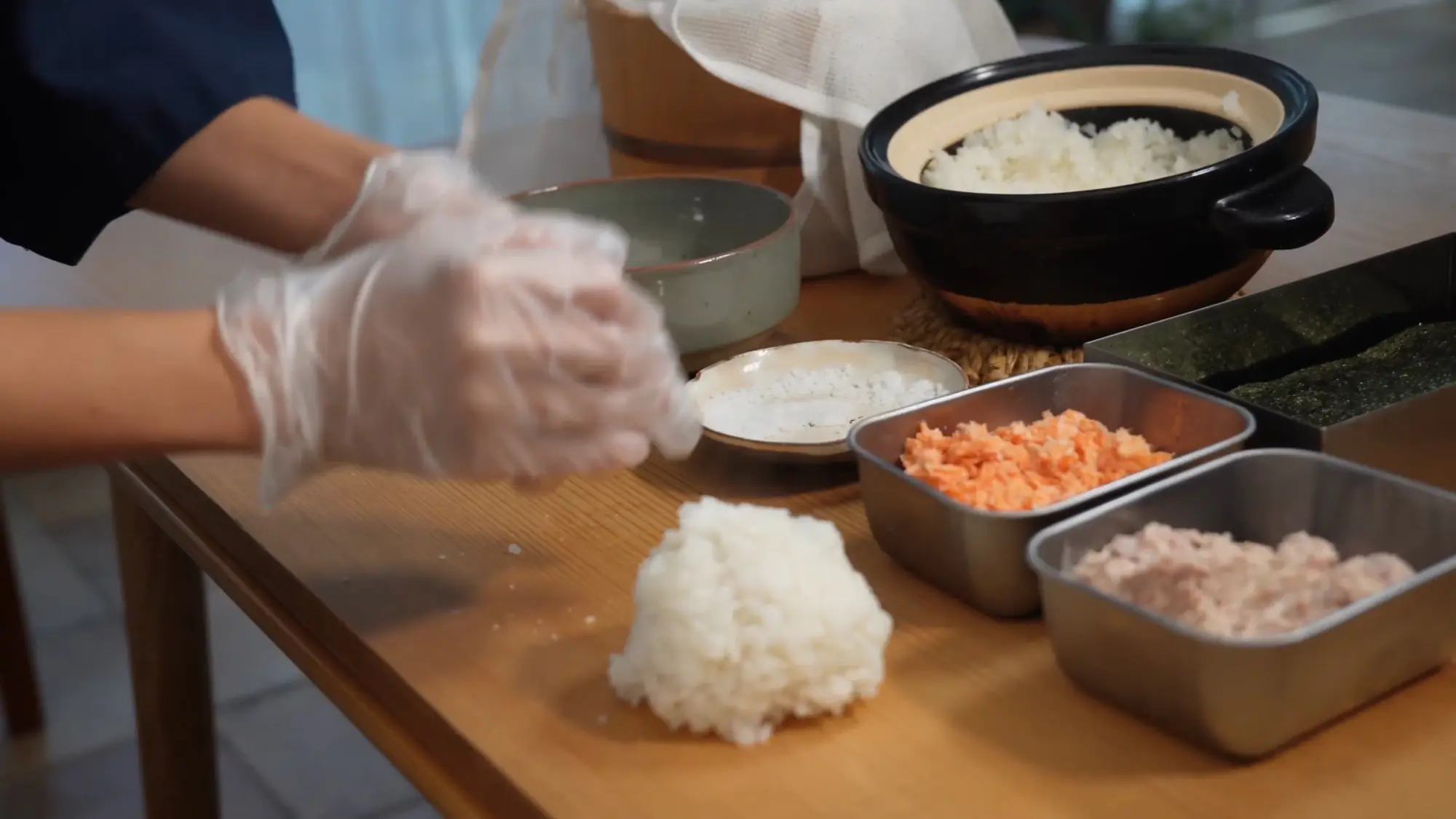 An onigiri-making station with a tray of toasted nori sheets, fresh rice, and trays of salmon and tuna fillings