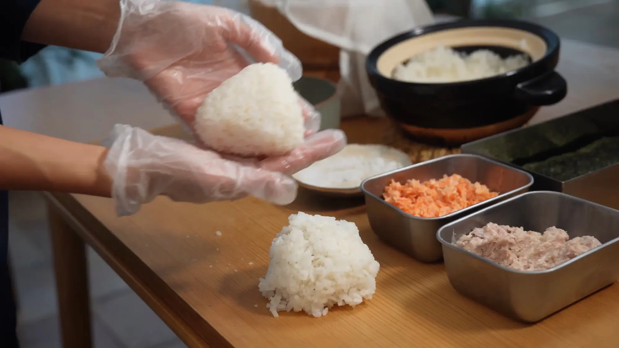 Gloved hands cupping a hand-pressed triangular rice ball, a second mound of rice resting on the board