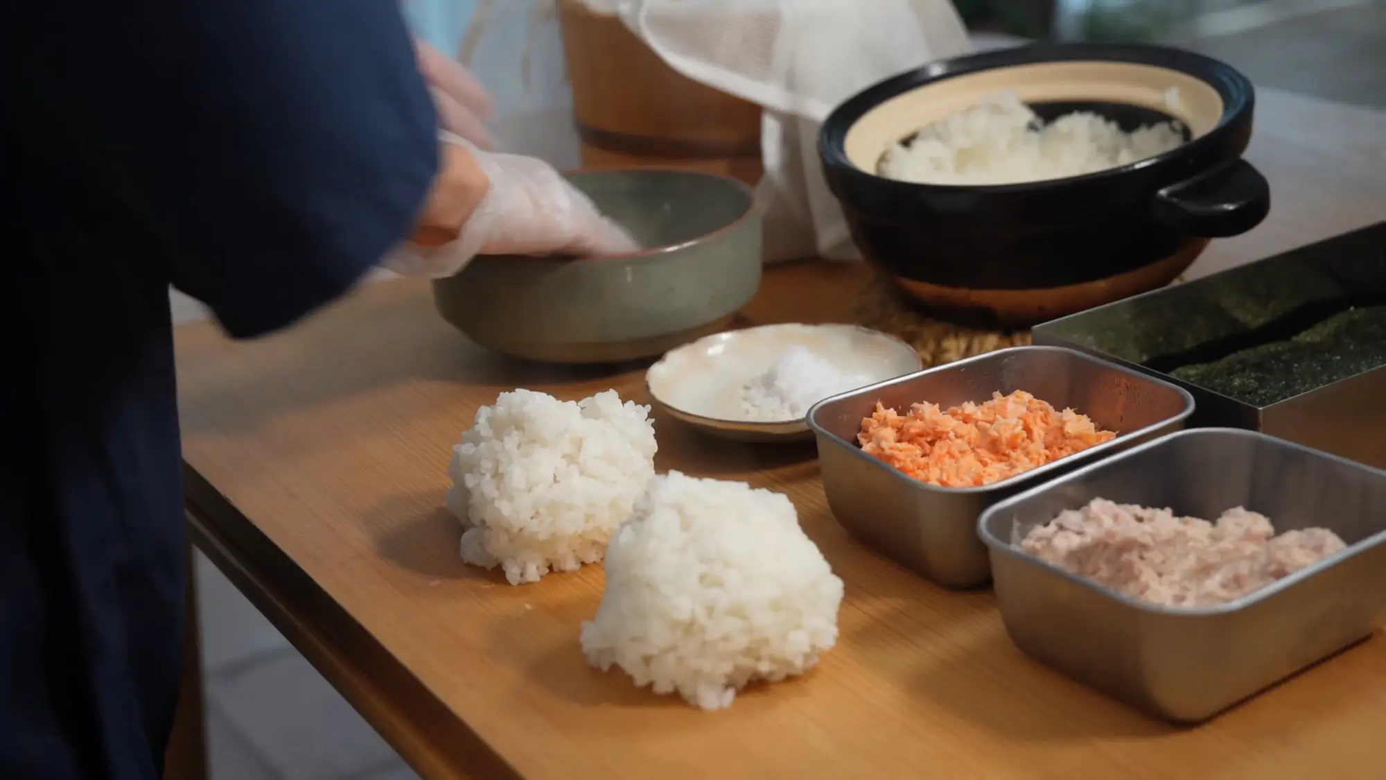 A full onigiri-making spread — hand-pressed rice balls, a dish of salt, trays of salmon and tuna, nori and a donabe of rice