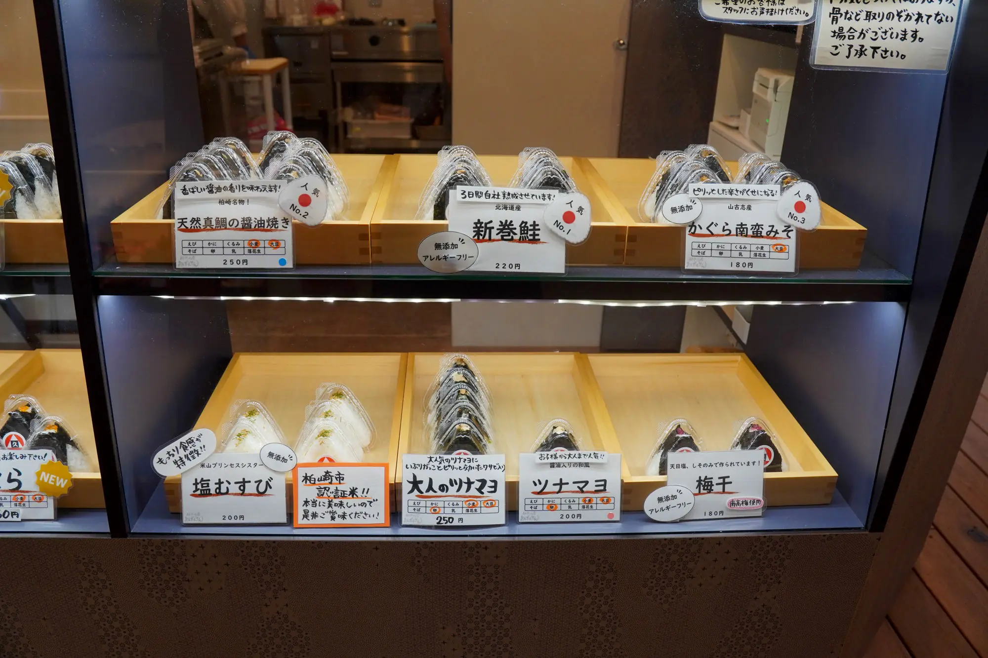 A glass display case showing onigiri varieties — sea bream shoyu, new-salted salmon, kagura nanban miso, shio-musubi, adult tuna mayo, tuna mayo, and umeboshi
